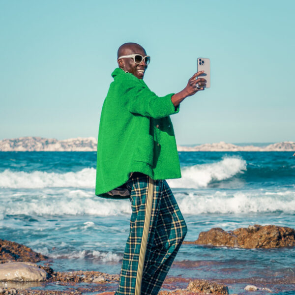Photographe Marketing - Personne stylée en manteau vert prenant un selfie sur une plage à Marseille, illustrant un style de vie moderne et connecté.