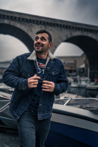 Tarifs 15 Portrait d’un homme souriant sous la pluie à Marseille, portant une veste en jean avec un port et un pont en arrière-plan.