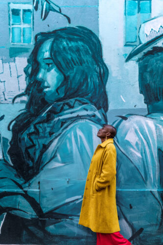 Tarifs 13 Portrait artistique en extérieur à Marseille, avec une personne en manteau jaune contemplant une fresque murale bleue représentant une femme.