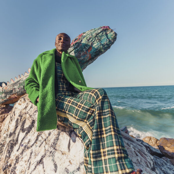 Photographe Portrait Marseille 3 Portrait stylé d'une personne vêtue d'un manteau vert et d'un pantalon à carreaux, assise sur un rocher en bord de mer à Marseille.